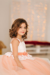 Little beautiful girl with brown hair in a Peach-colored dress. Girl sitting on the sofa