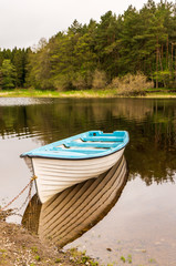 Naklejka na ściany i meble White fishing boat tied and anchored at Blessington Lake shore in Wicklow, Ireland, reflected in the calm water, on a beautiful forest background. Peaceful morning landscape,