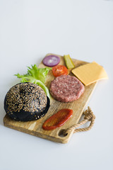 Ingredients for Burger, black bun, green salad, onion, tomato, cucumber, cheese, beef steak, sauce on wooden chopping Board, White background, side view