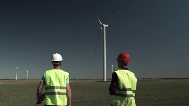 Two Young Engineers Men Approach A Windmill Power Generator And Discuss The Plan Of Its Repair In The Field On A Sunny, Clear Day