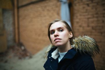 Fototapeta premium Hipster girl wearing dark blue jacket, gray sweater and jeans posing against rough street wall, minimalist urban clothing style