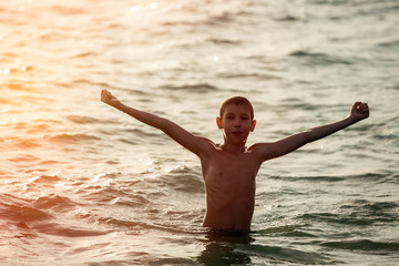 Silhouette of happy little boy jumping in sea with hands up at sunset