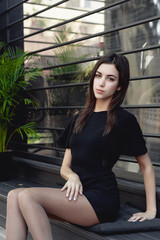 portrait of a young beautiful brunette woman on the street, model posing, women's urban street fashion, strict sexy black dress