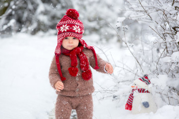 Portrait of a cute toddler baby dressed in a brown hand knitted jacket, pants, red hat and scarf,...