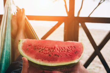 Young man holding piece of juicy watermelon in hands. He lying on hammock at sunny beach by ocean.