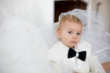 Portrait of elegant handsome little boy in a tuxedo, lying in bed on his moms bridal dress