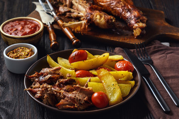 Roasted turkey legs with potatoes and tomatoes, on dark wooden background.