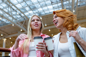 Two beautiful smiling women with disposable coffee cups talking in shopping center. Blur mall as background. Lifestyle and friendship concepts.