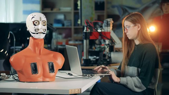 Human-like robot is moving its mouth under control of a young woman