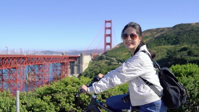 young girl tourist carrying backpack and camera joining cycle tour in san francisco stop and seeing view of golden gate bridge from high mountain spot point on sunny day. female traveler riding bike.
