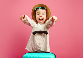 Little girl with a suitcase expresses surprise and shock, opened her mouth wide, wears beach hat and sunglasses, pointing fingers at you, stands on pink isolated background.