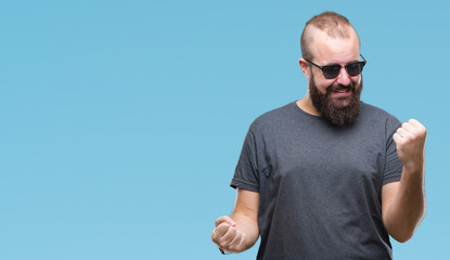 Naklejka premium Young caucasian hipster man wearing sunglasses over isolated background celebrating surprised and amazed for success with arms raised and open eyes. Winner concept.