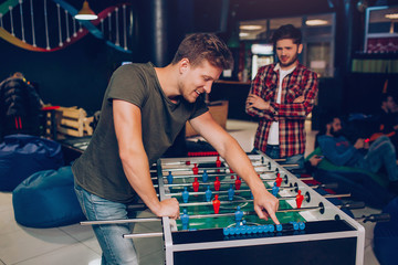 Happy young man stand and lean at table soccer in playing room. His unhappy friend stand behind. He hold hands crossed.
