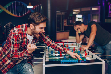 Youn bearded happy man cheering at soccer table in playing room. He wins. Another guy stand behind....