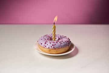 burning candle in middle of doughnut with sprinkles on white table on pink background
