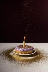 burning candle in middle of tasty donut with falling sparkles isolated on black