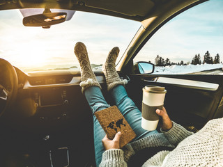 Woman drinking coffee paper cup inside car with feet warm socks on dashboard