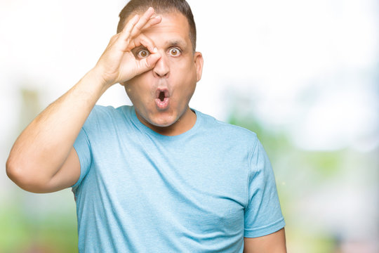 Middle age arab man wearing blue t-shirt over isolated background doing ok gesture shocked with surprised face, eye looking through fingers. Unbelieving expression.