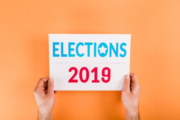 cropped view of man holding card with 'elections 2019' lettering isolated on orange