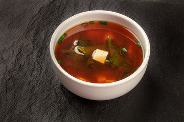 A photo of a bowl of miso soup with tofu, scallions, and wakame algae, on a black background with a place for text