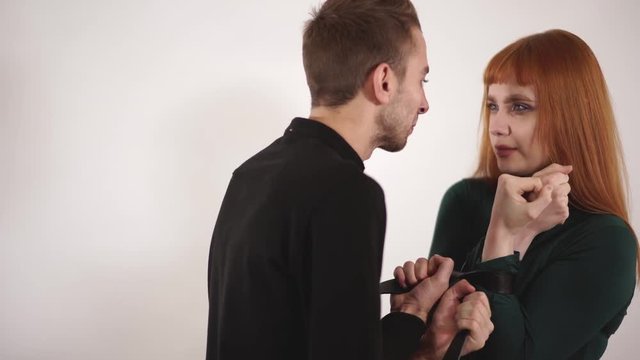 Aggressive angry man yelling at crying young woman with red long hair and tie females hands with belt showing his violation.