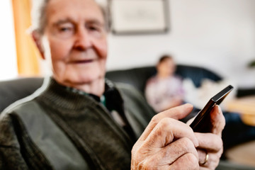 Obraz na płótnie Canvas Senior sitzt zuhause im Wohnzimmer und surft auf seinem smart phone im Internet