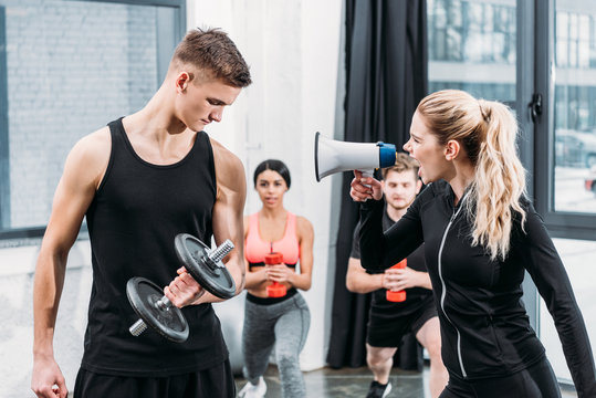 sportswoman with megaphone yelling at young man training with dumbbell in gym