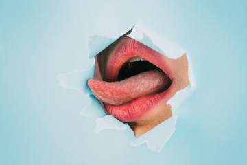 Woman's open mouth with sexy pink lips and tongue. Fashion photography. Natural full lips with bright lip makeup. Pink lipstick and lip gloss in a paper hole, blue background.