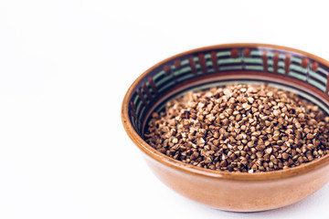 Dry raw buckwheat grains in brown ceramic bowl on white wooden background.