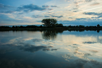 Clouds reflecting in the water of a calm lake, the horizon and a group of trees on the shore