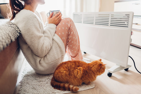 Using heater at home in winter. Woman warming and drinking tea with cat. Heating season.