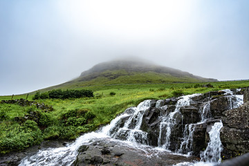 Fototapeta premium Beautiful and peaceful waterfall (long exposure) with green grass and small houses in foggy weather in Bour village, Faroe Islands, North Europe travel destination