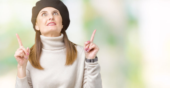 Middle age mature woman wearing winter sweater and beret over isolated background amazed and surprised looking up and pointing with fingers and raised arms.