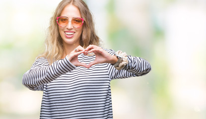 Beautiful young blonde woman wearing sunglasses over isolated background smiling in love showing heart symbol and shape with hands. Romantic concept.
