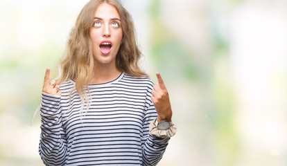 Beautiful young blonde woman wearing stripes sweater over isolated background amazed and surprised looking up and pointing with fingers and raised arms.