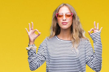 Beautiful young blonde woman wearing sunglasses over isolated background relax and smiling with eyes closed doing meditation gesture with fingers. Yoga concept.