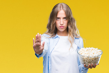 Beautiful young blonde woman eating popcorn over isolated background with open hand doing stop sign with serious and confident expression, defense gesture