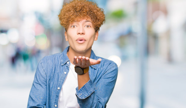 Young handsome man with afro hair wearing denim jacket looking at the camera blowing a kiss with hand on air being lovely and sexy. Love expression.