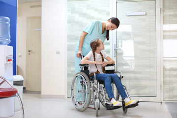 Young female doctor taking care of little girl in wheelchair indoors
