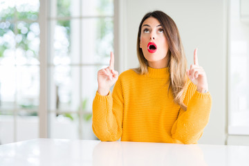 Young beautiful woman wearing winter sweater at home amazed and surprised looking up and pointing with fingers and raised arms.