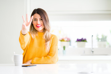 Young beautiful woman drinking a cup of coffee at home showing and pointing up with fingers number three while smiling confident and happy.