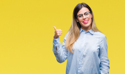 Young beautiful business woman wearing glasses over isolated background smiling with happy face looking and pointing to the side with thumb up.