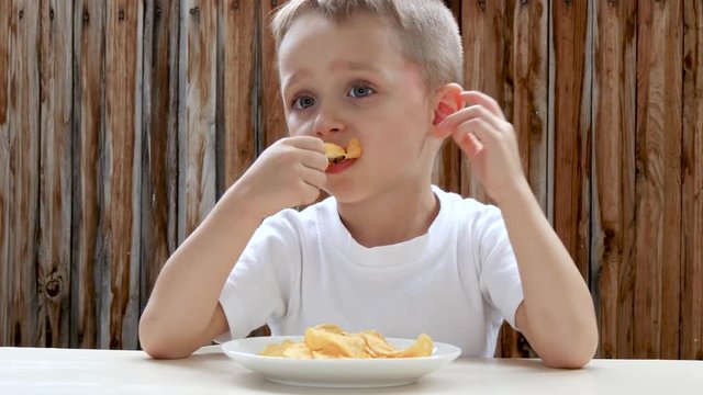 A child eats potato chips with pleasure against the background of a wooden wall. The boy is pleased with the food and shows a thumb up.