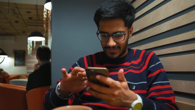 Indian Guy Scans the News on the Smartphone and Smiles While Sitting at the Cafe