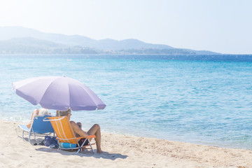 Man and woman sitting on small fold-out chairs on the beach under purple sunshade with a view of the blue sea water
