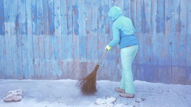 Slender woman in a blue winter ski suit sweeps snow in courtyard of village house after heavy snowfall. Big broom.
