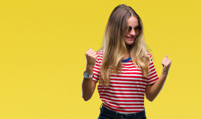 Young beautiful blonde woman wearing sunglasses over isolated background very happy and excited doing winner gesture with arms raised, smiling and screaming for success. Celebration concept.