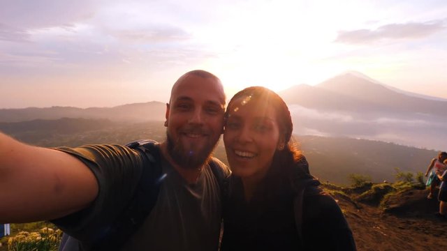 Young Mixed Race Couple Taking Selfie Portrait on Top of Batur Volcano with Beautiful Sunrise Landscape on Background. 4K Slowmotion Lifestyle Hiking Travel Concept Footage. Bali, Indonesia.