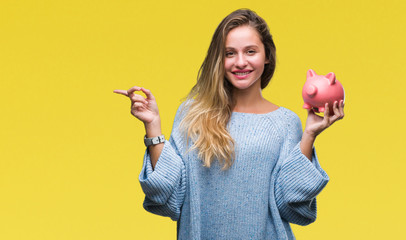 Young beautiful blonde woman holding piggy bank over isolated background very happy pointing with...
