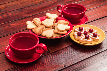 Heart-shaped biscuits with cream and frozen cherries, two cups of tea in bright red cups on a tinted wooden table top